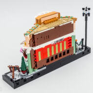 Miniature de bâtiment en briques LEGO de style festif, avec un panneau décoratif coloré et une lampe de rue, sur une plaque de base noire.