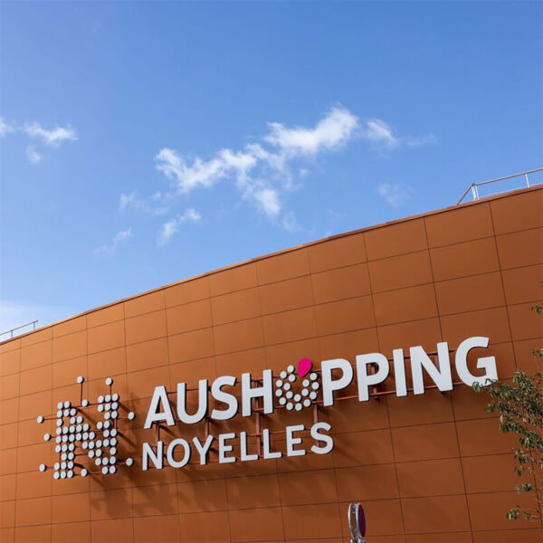 Aushopping Noyelles façade orange avec logo lumineux moderne, centre commercial dynamique, architecture innovante, shopping, boutiques, loisirs, aire commerciale, design contemporain, signalétique visible.
