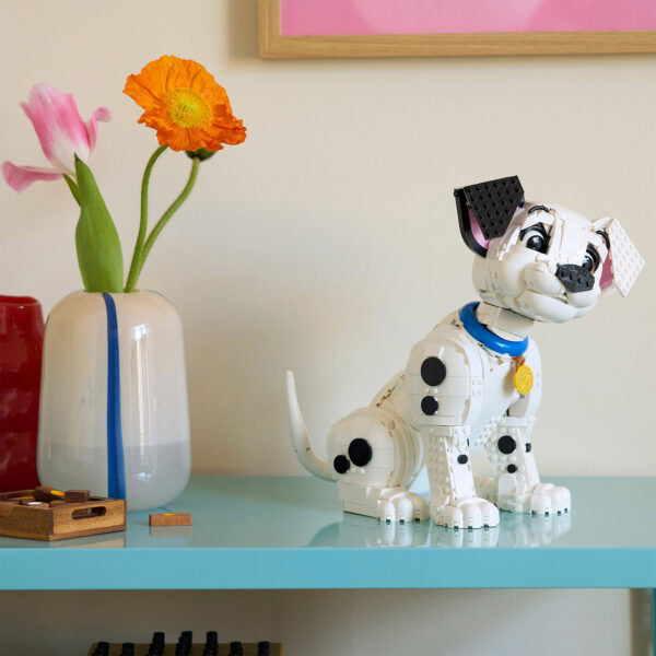Lego chien robotique en briques blanches avec collier bleu et médaillon or, décoration intérieure moderne avec vase de fleurs colorées sur une table bleu pastel.