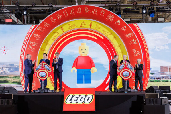 LEGO lors d'une cérémonie de lancement avec une grande sculpture LEGO, des personnes en costume, un décor coloré, et un logo LEGO visible, soulignant l'innovation et la créativité LEGO.
